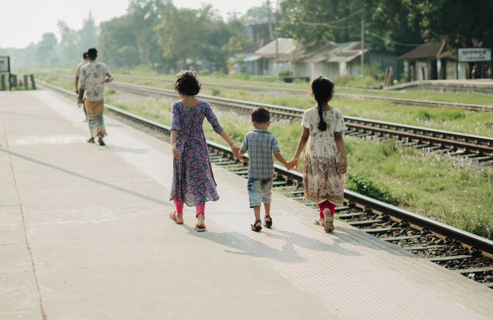 Three children holding hands as they walk along a sunny railway platform.