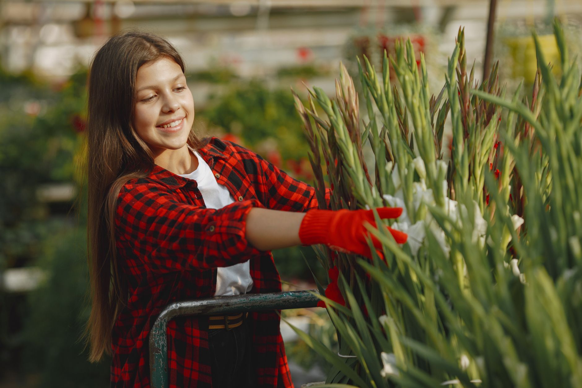 Smiling girl in a greenhouse tending to plants, wearing red plaid shirt and gloves.
