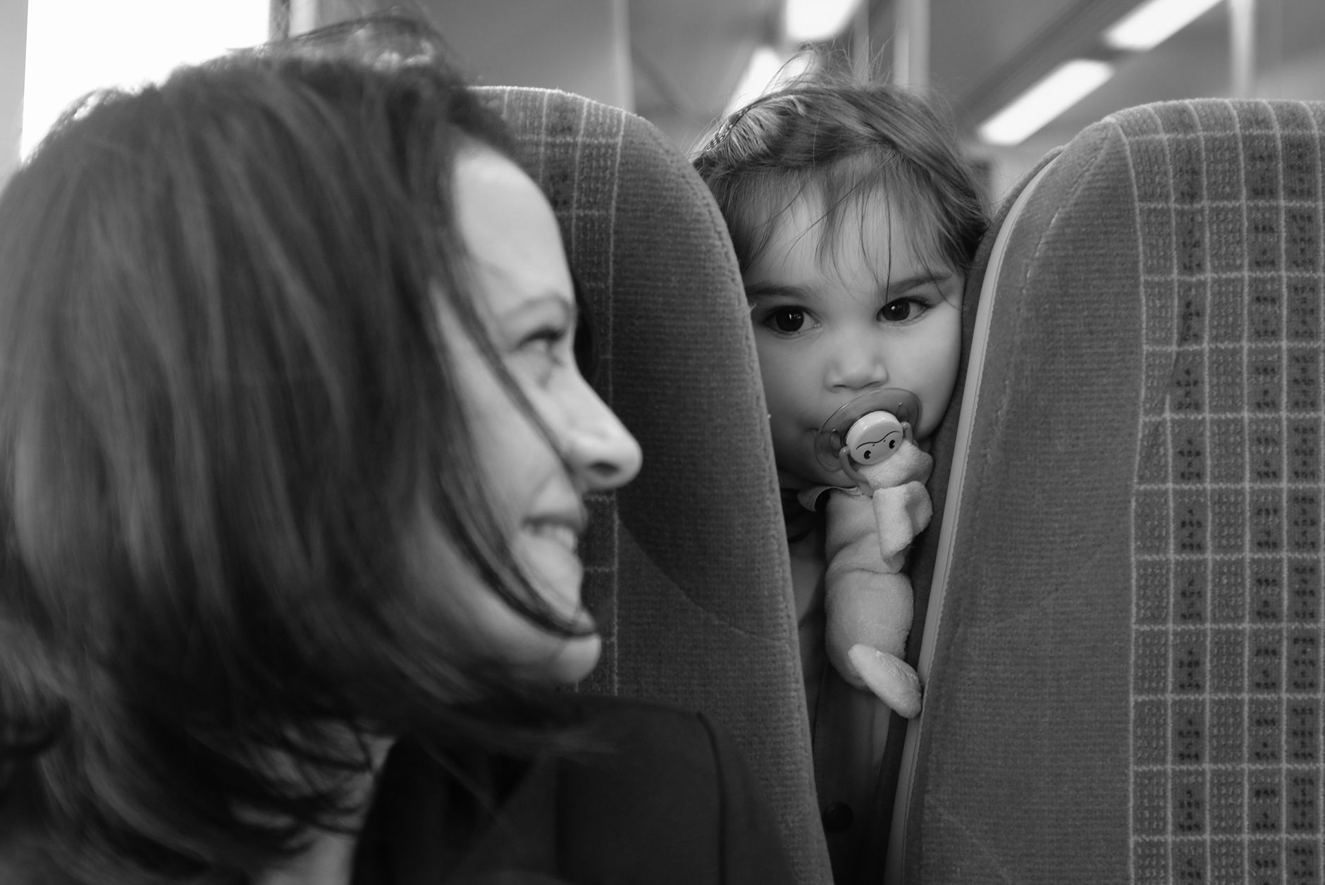 Mother smiling at her child playing peekaboo on a train, captured in black and white.
