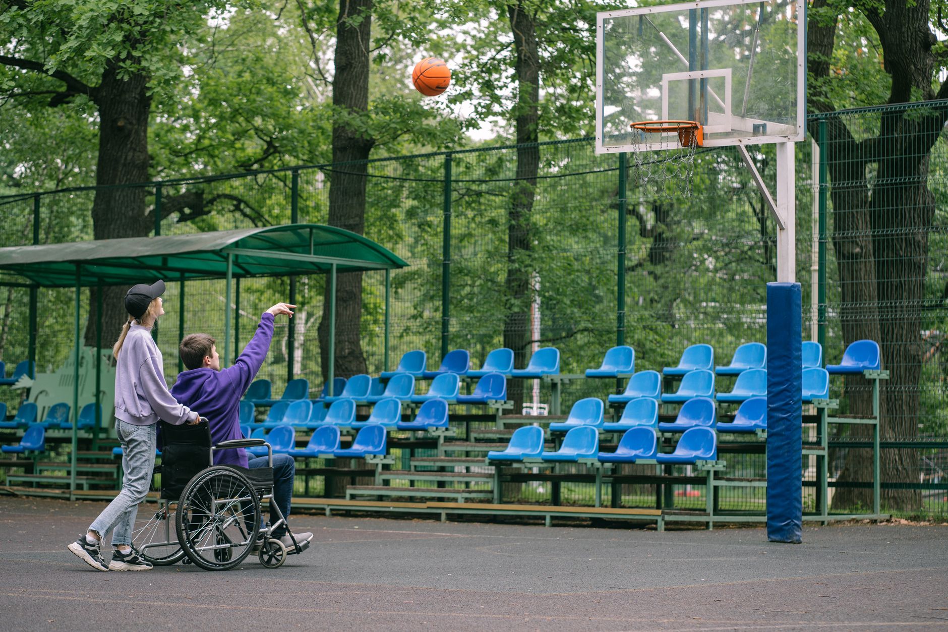 A person in a wheelchair playing basketball with a friend outdoors on a court.