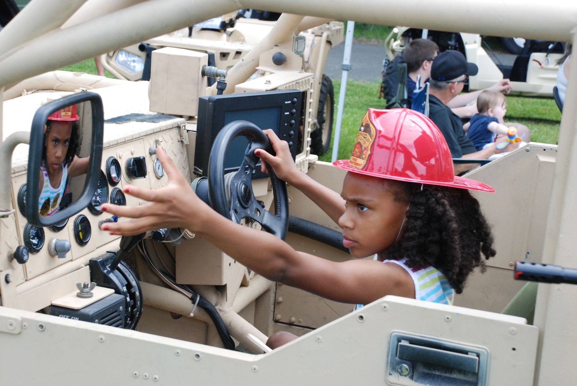 Young girl wearing a firefighter hat in a military jeep, pretending to drive.