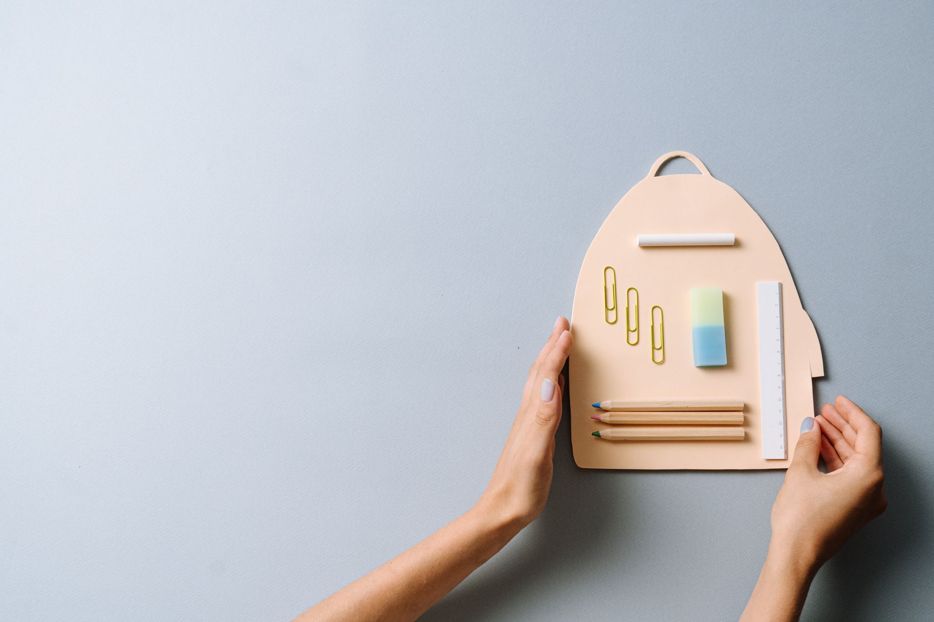 Flat lay of school supplies on a cutout in backpack shape, held by hands on a light surface.