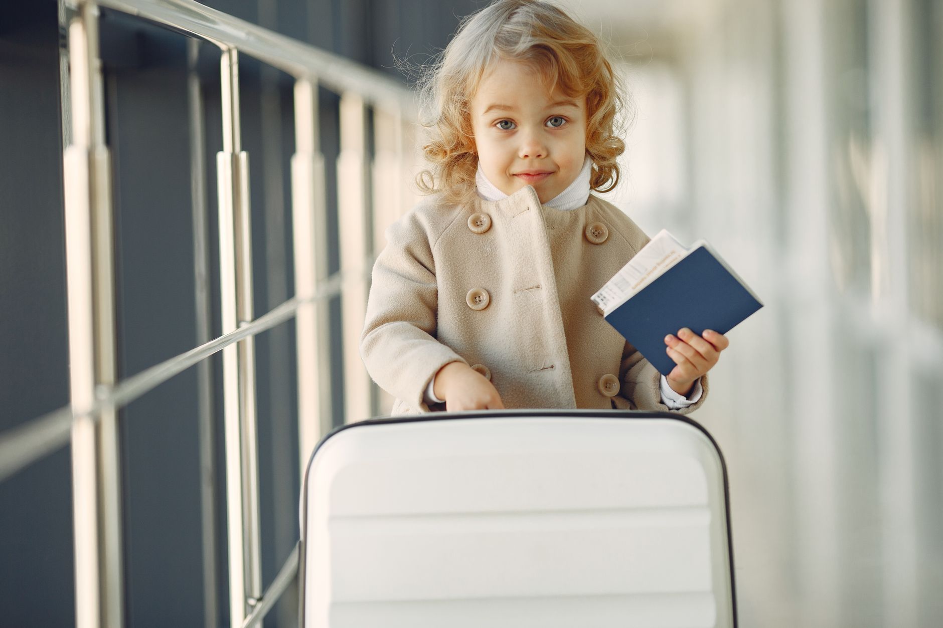 Adorable little girl with blond curly hair in stylish beige coat standing with white suitcase and passport in airport corridor