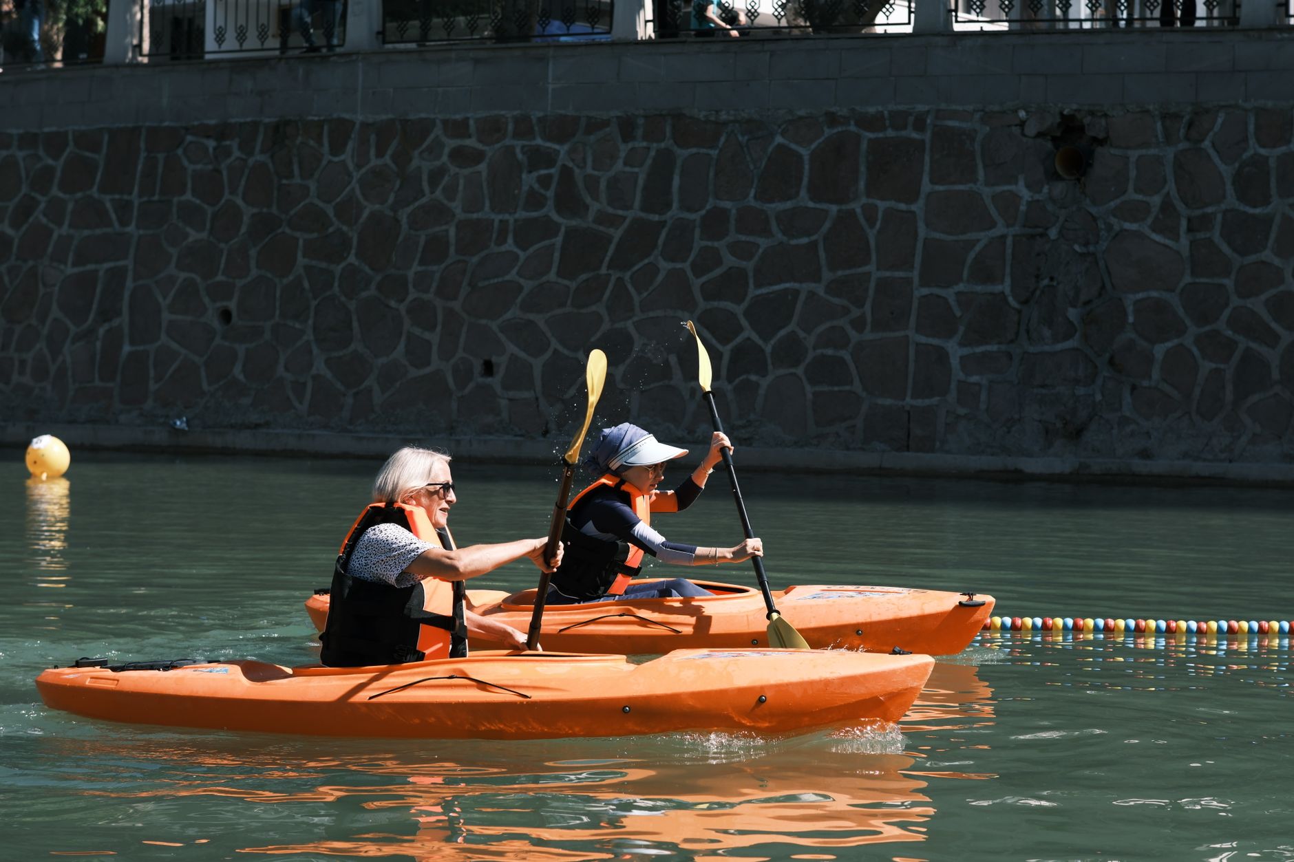 Two people kayaking in orange boats on a sunny day by stone wall.