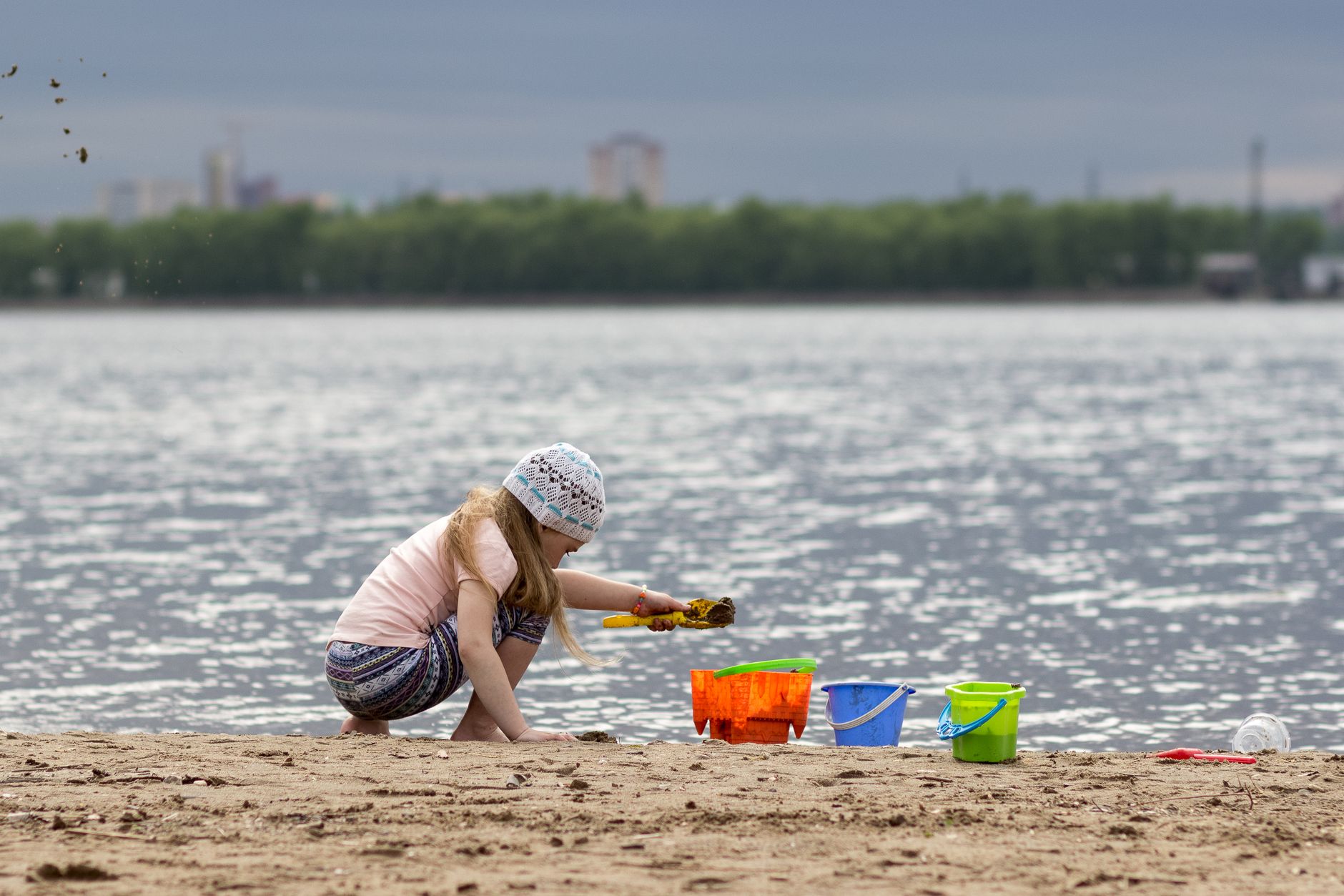A young girl plays with colorful buckets on a sandy beach by a lake on a summer day.