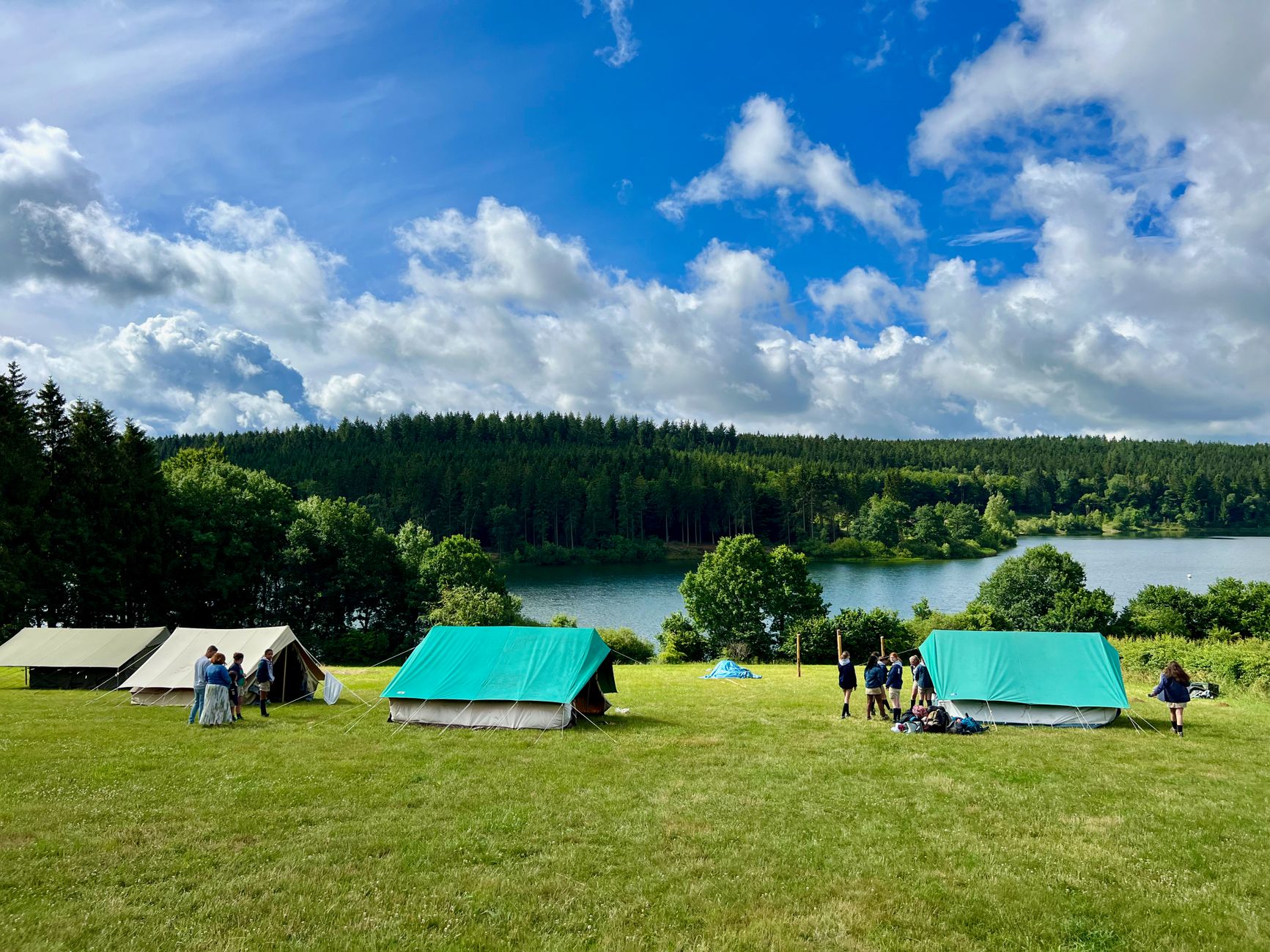 A vibrant lakeside camping scene with tents, people, and lush greenery under a bright blue sky.