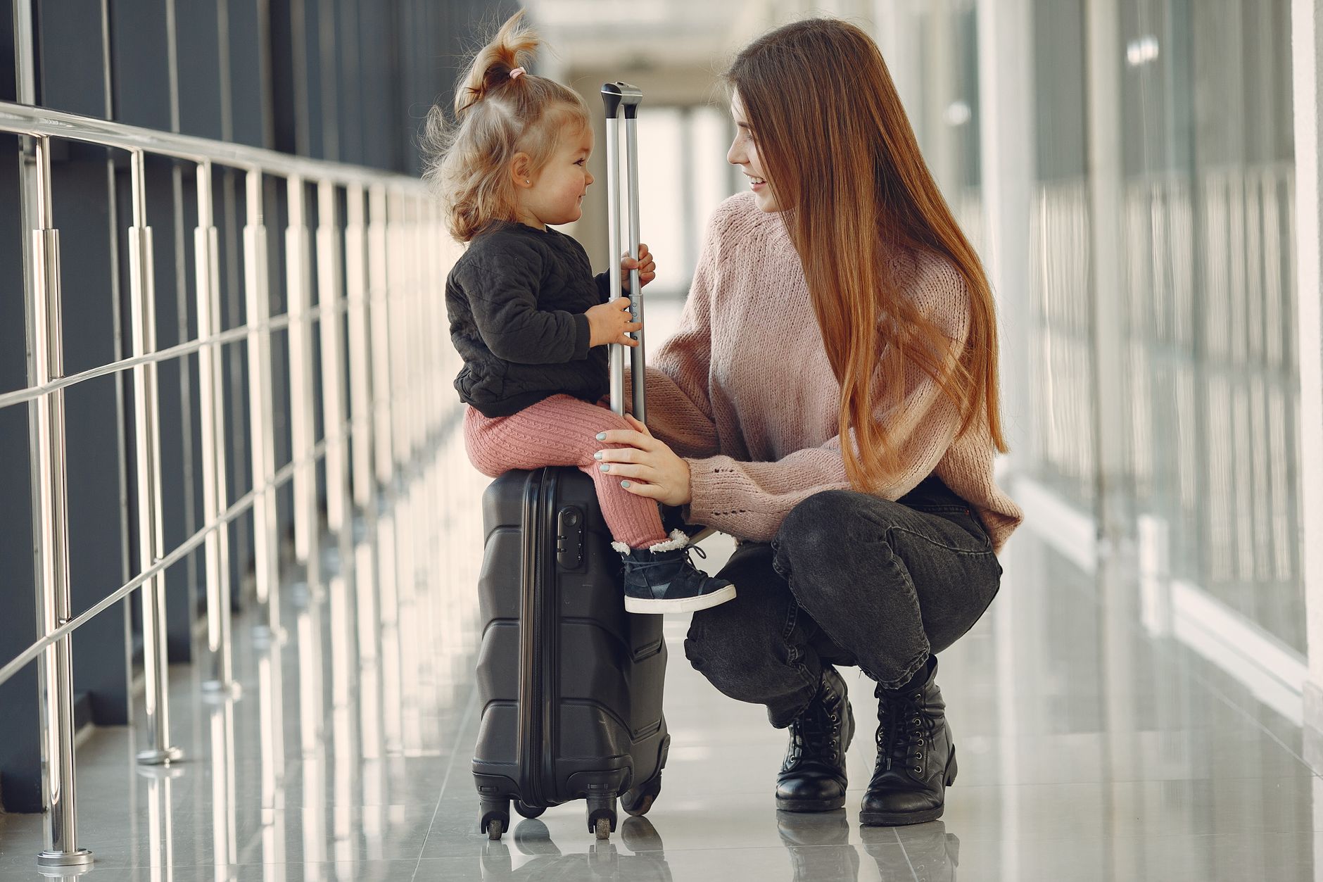 Full length of positive content woman and cute stylish blond little girl sitting on black suitcase in airport corridor while waiting for fli