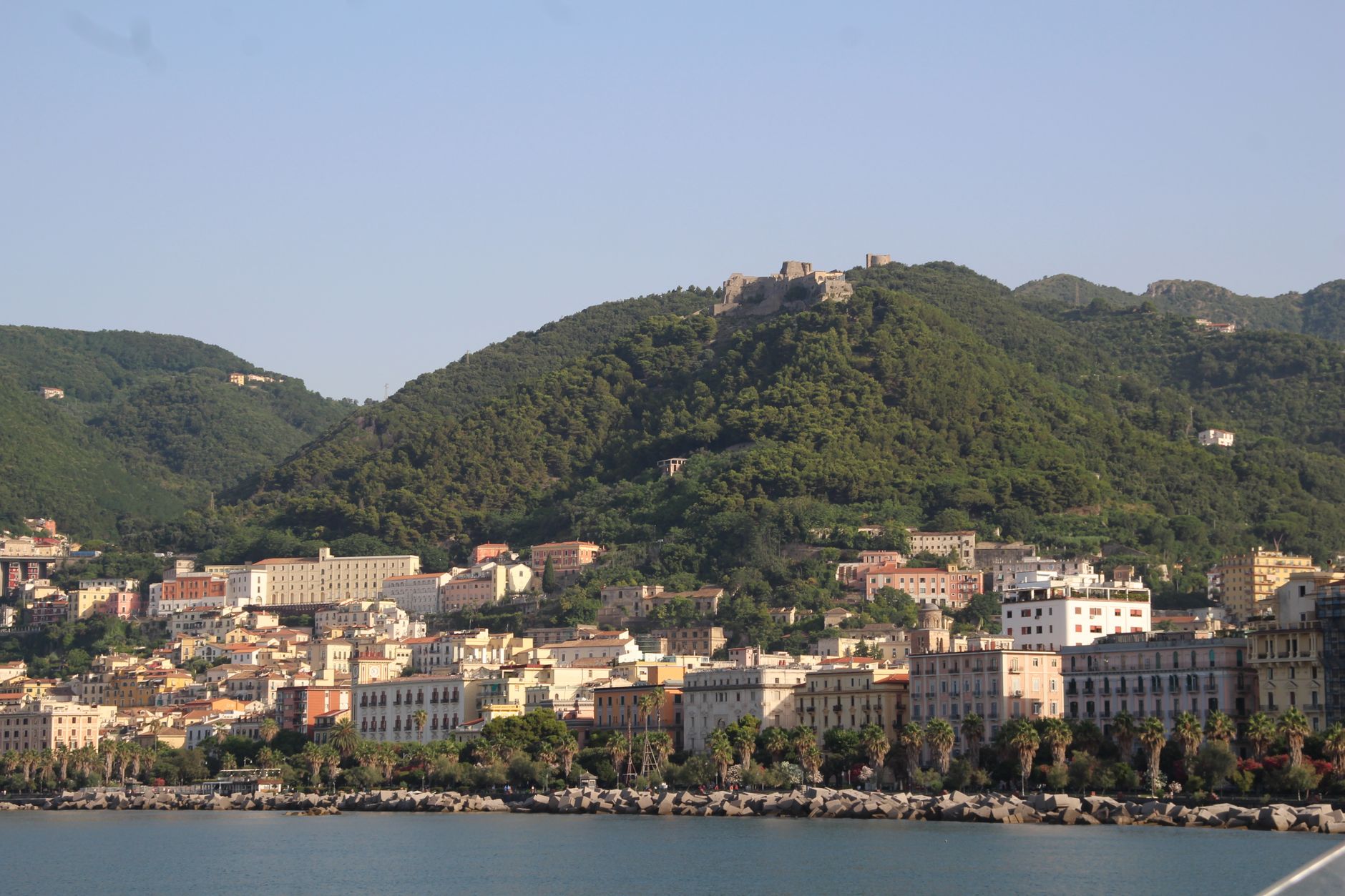 Panoramic view of Salerno's coastal cityscape flanked by lush green hills under a clear sky.