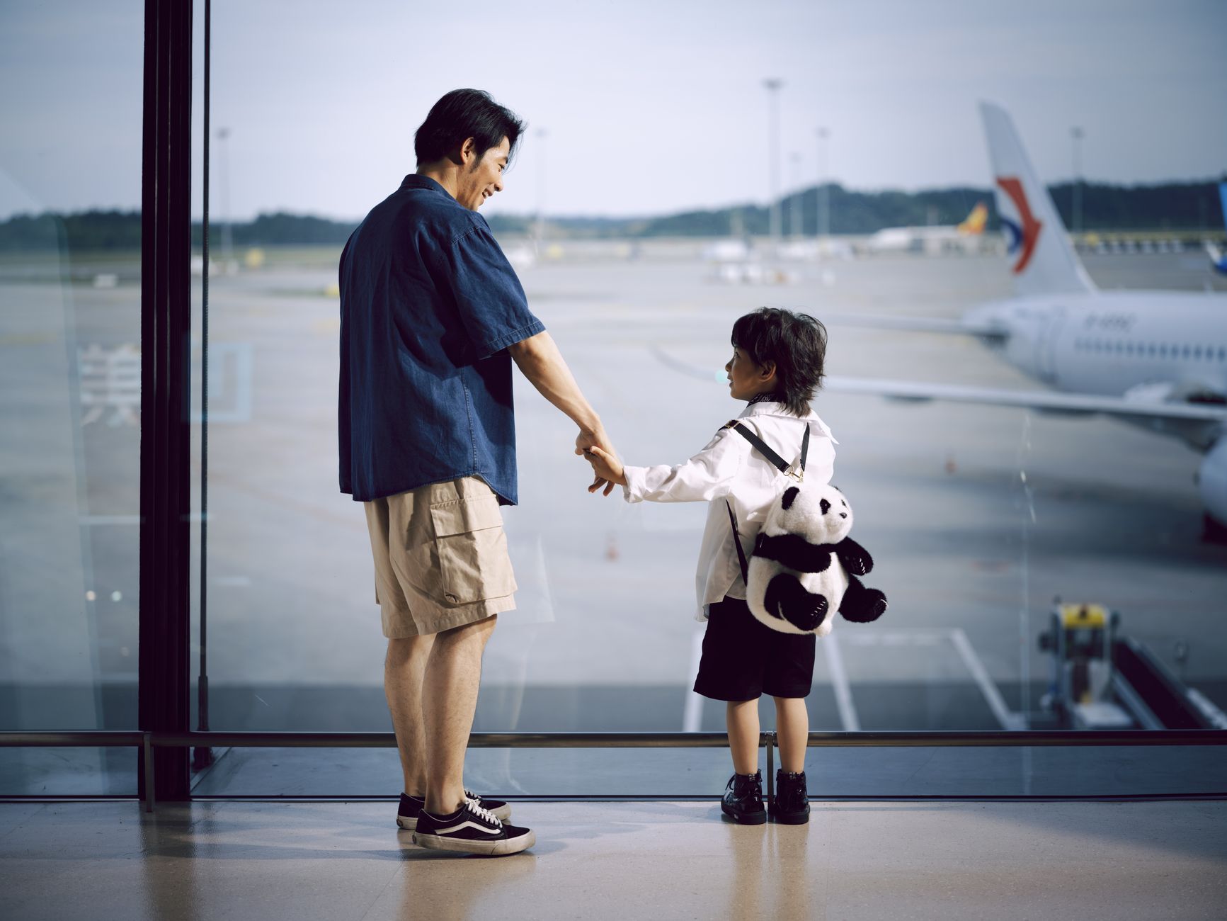 A father and child holding hands, gazing at planes through an airport window.