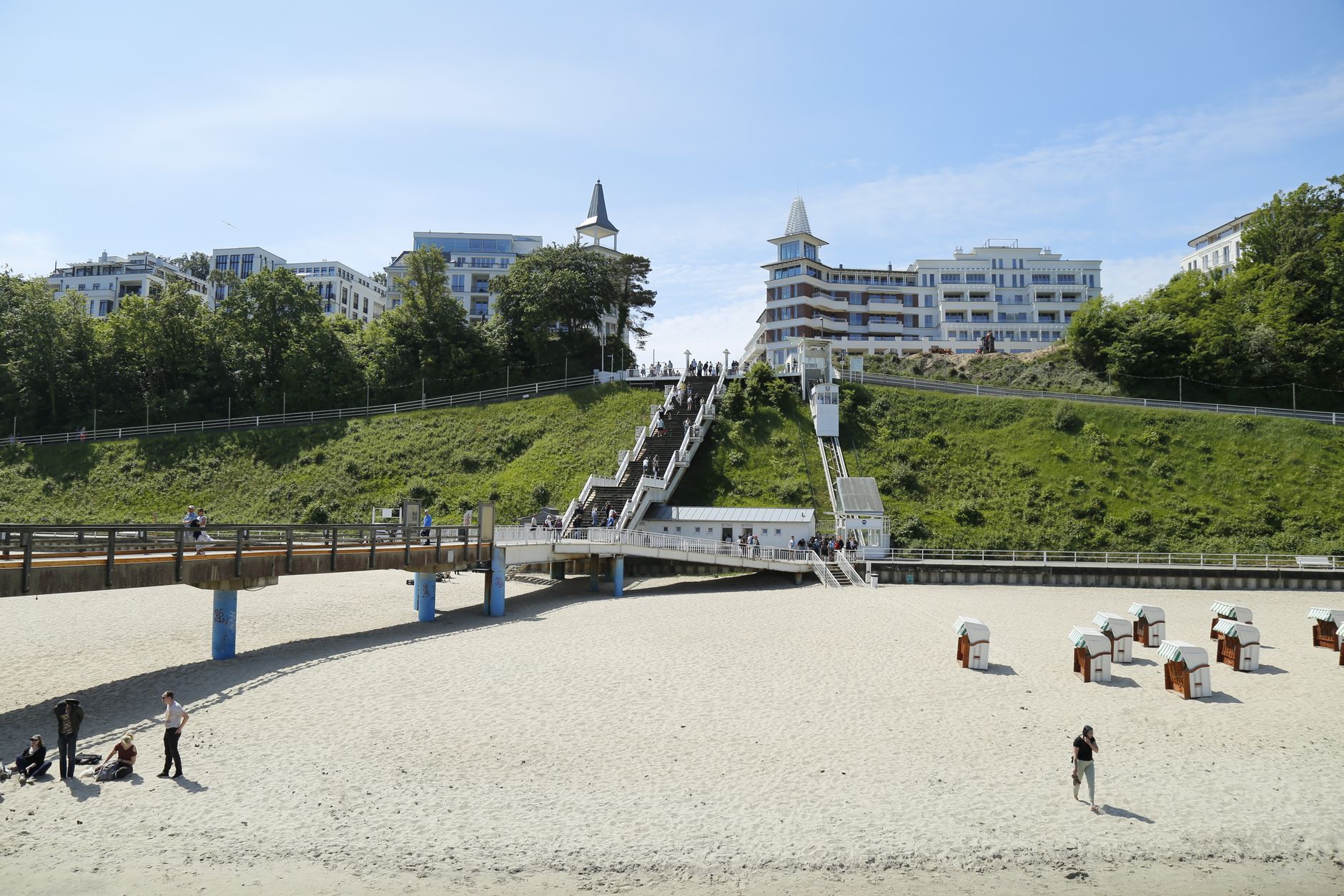 Beachfront scene showcasing modern architecture, lush greenery, and sandy shore with lounging tourists.
