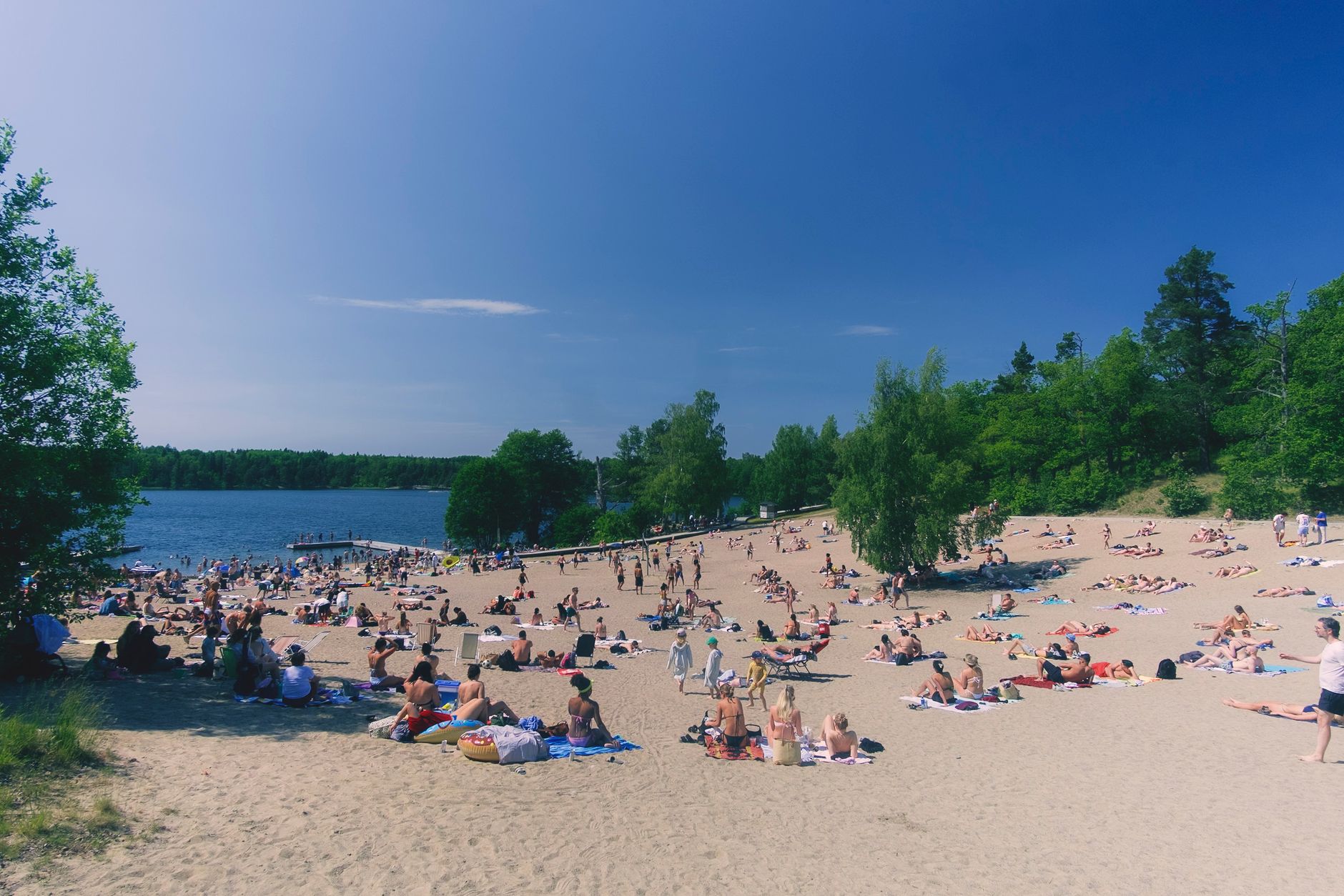 Crowd of people enjoying a sunny day at a sandy lakeside beach surrounded by lush greenery.