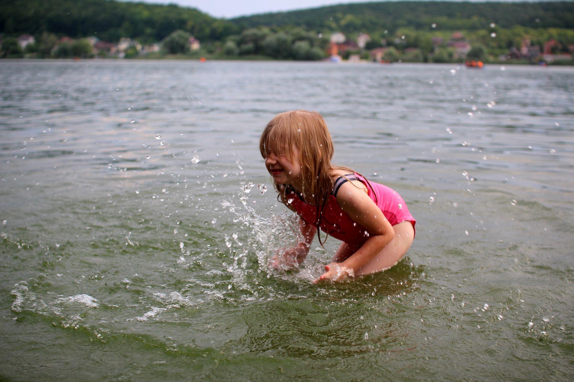 Smiling child splashing in the lake, enjoying a sunny day outdoors in a pink swimsuit.