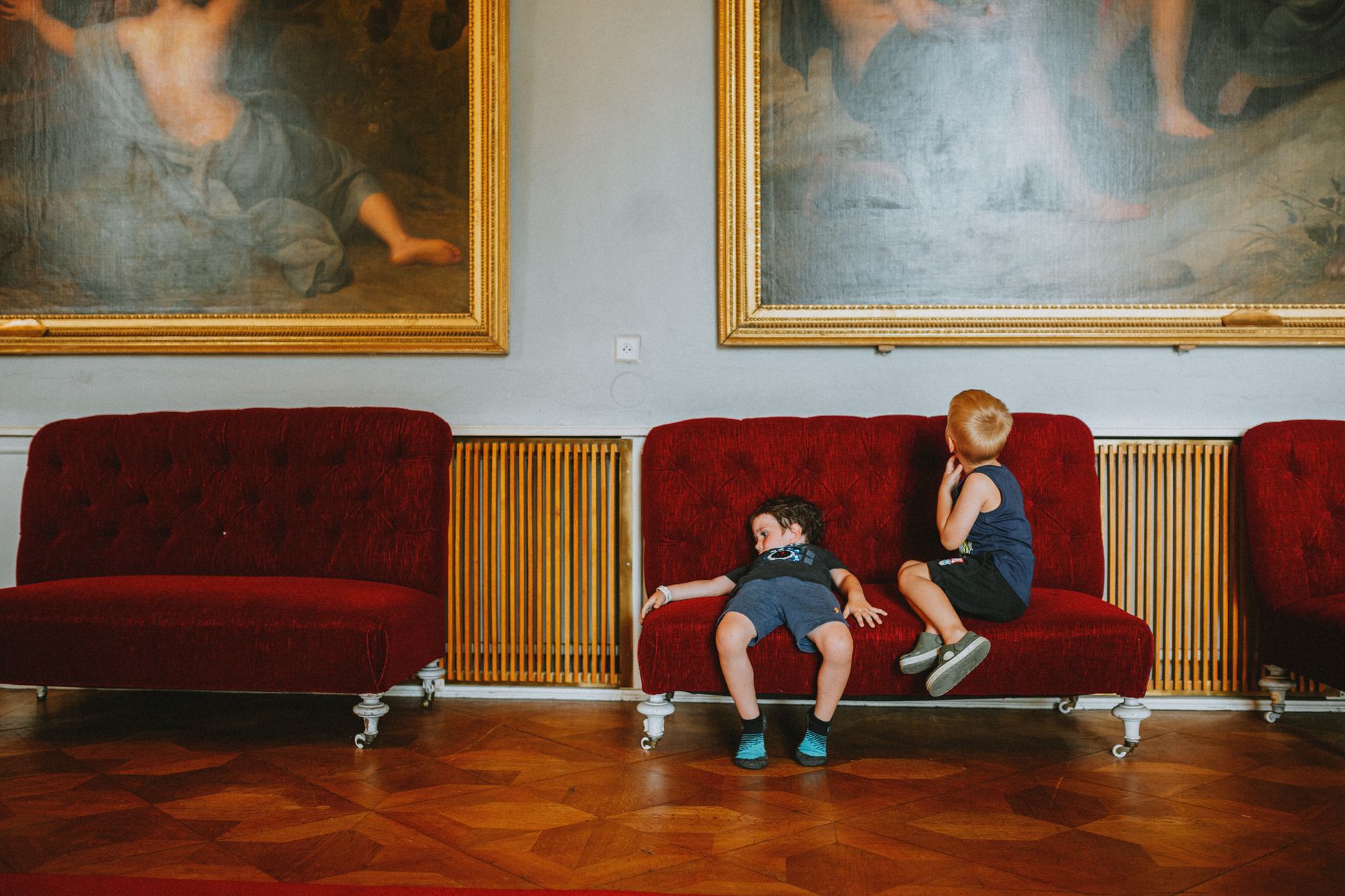 Two children sit on a red sofa in a museum, surrounded by vintage paintings.