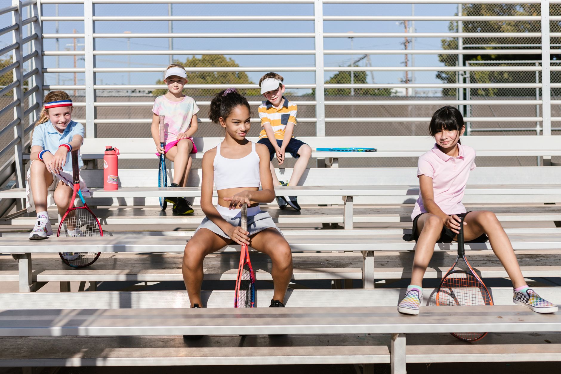 Group of children in sportswear sitting with tennis rackets on bleachers enjoying a sunny day.