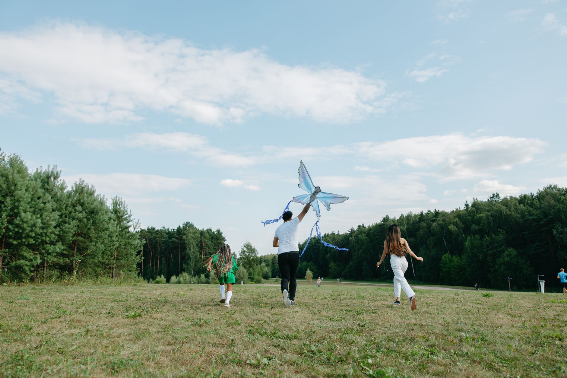 A family runs joyfully while flying a kite in a sunny park setting.