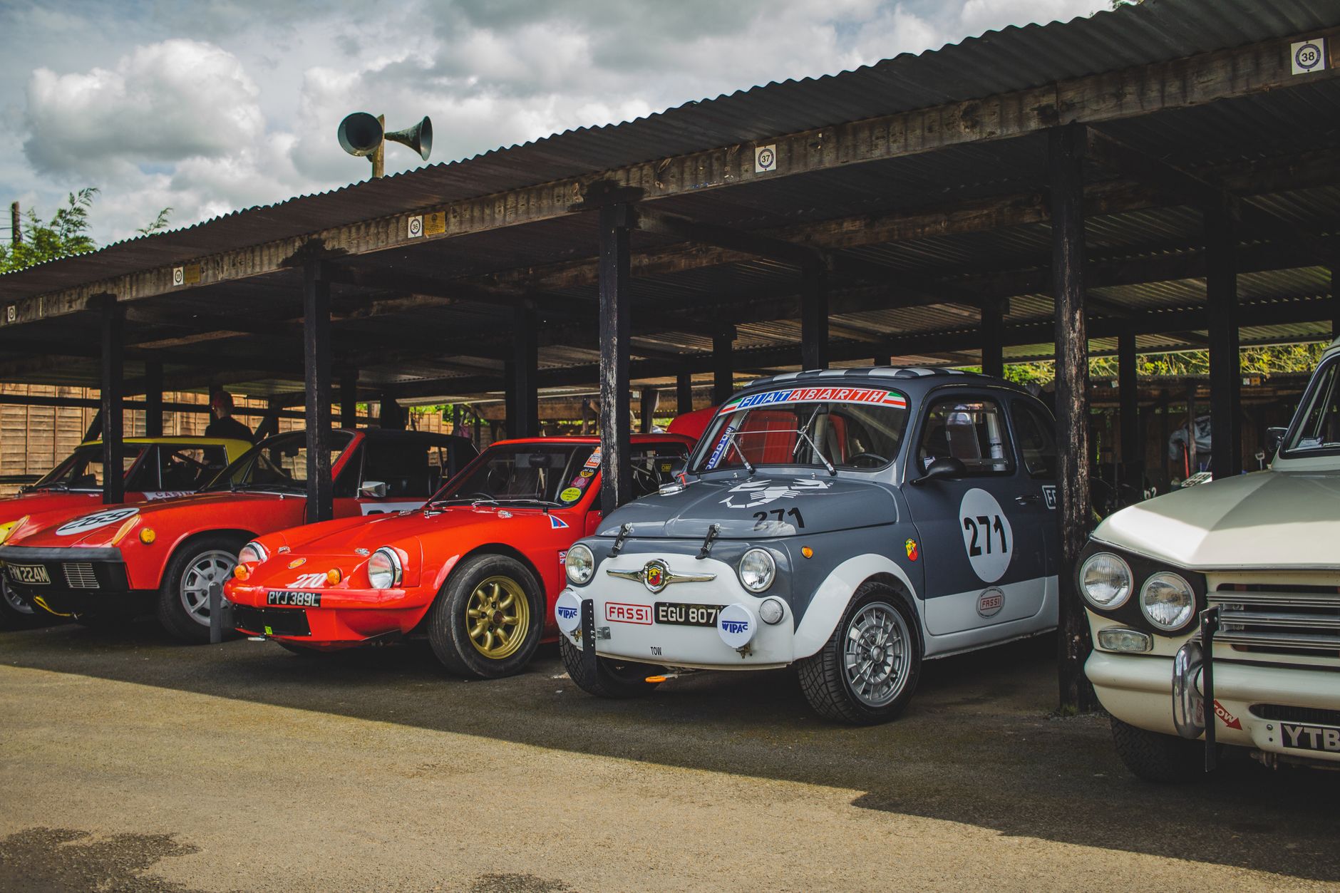 A collection of classic vintage cars parked under a wooden roof in an outdoor parking area.