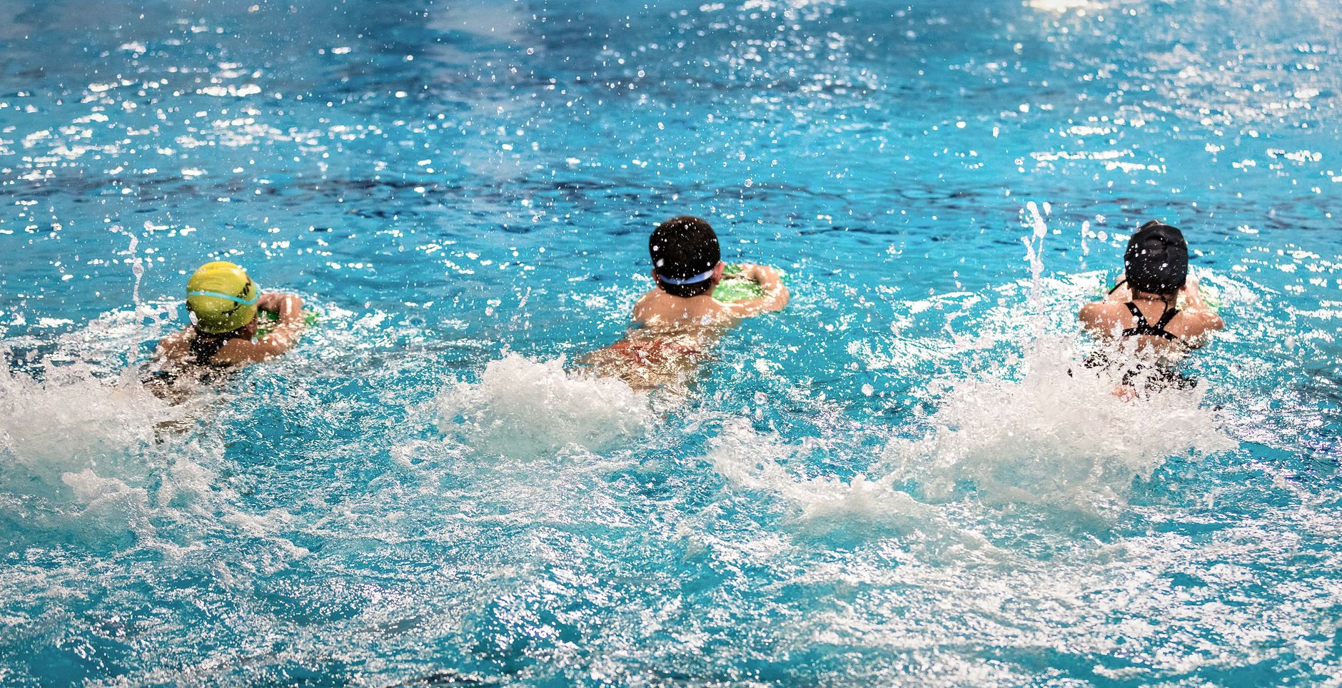 Three children swim freestyle in an indoor pool, splashing water, and having fun.