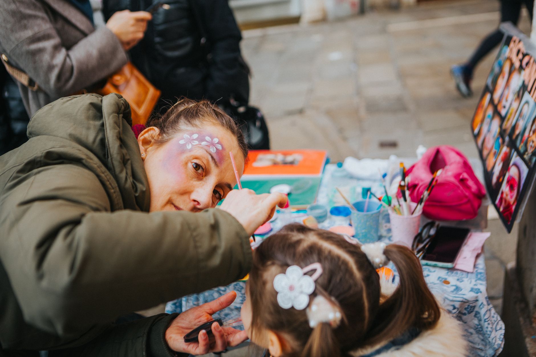 A face painter artistically decorates a child's face outdoors at a vibrant street festival.