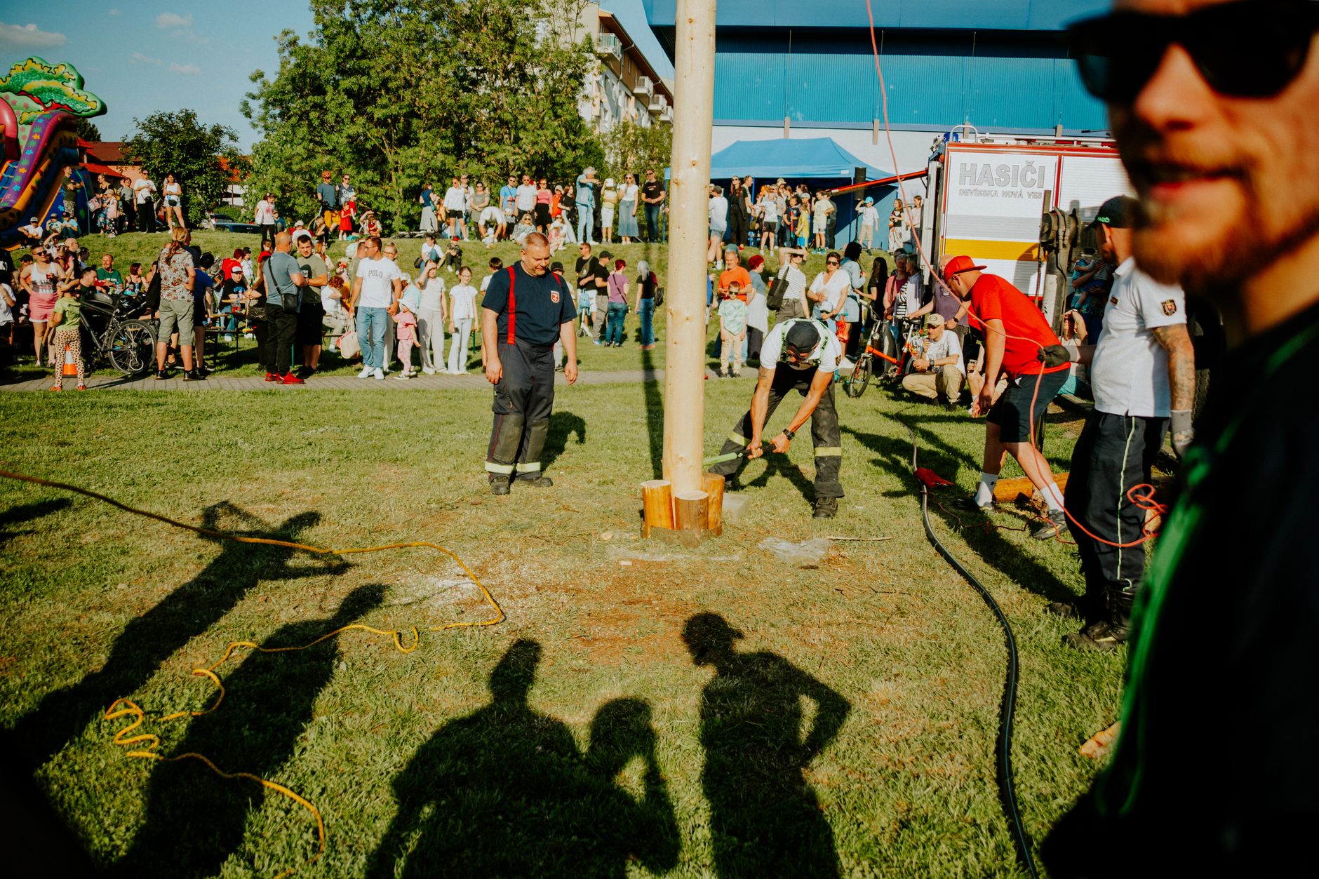 A vibrant summer festival with people gathered around a pole-raising event outdoors.