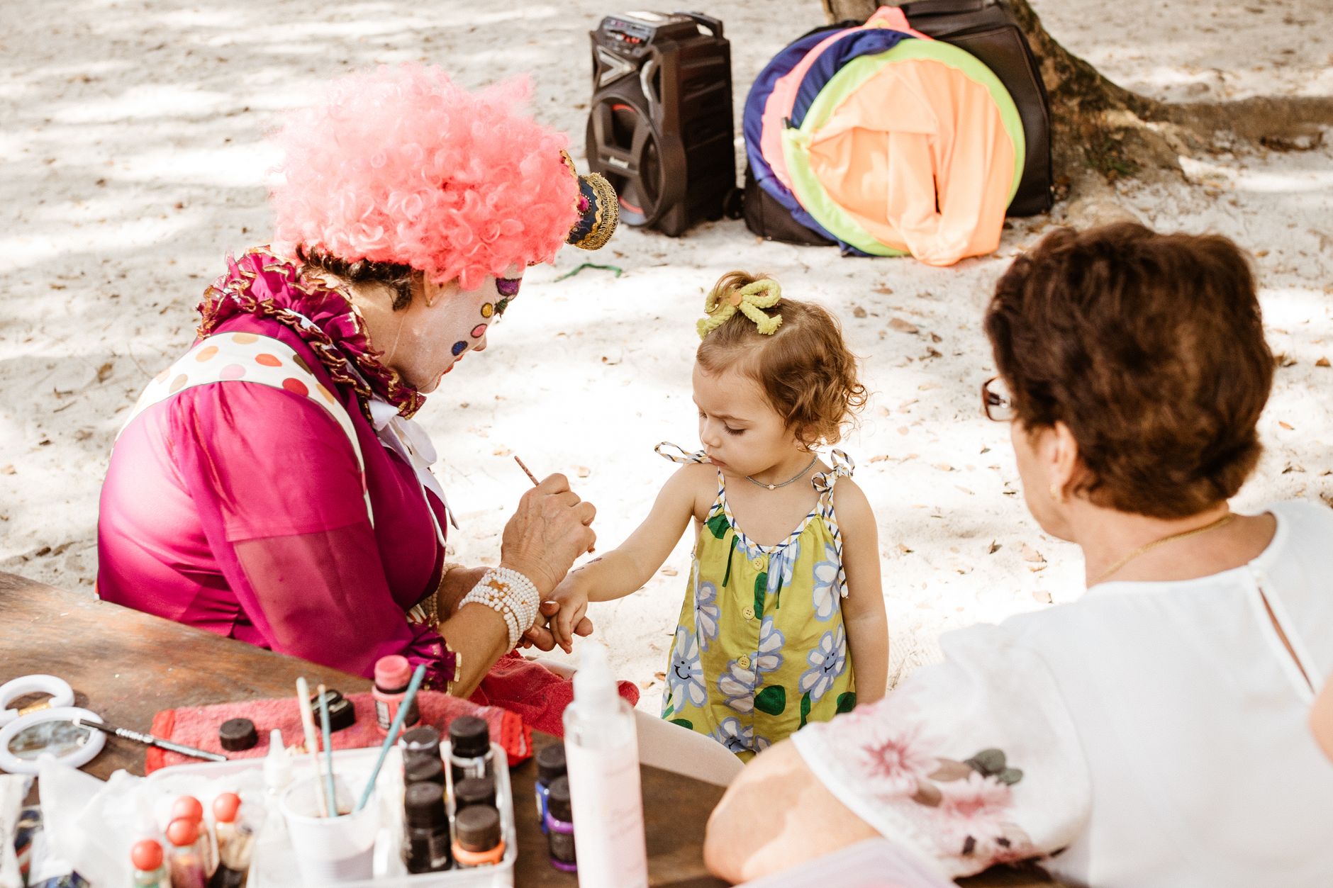 Vibrant outdoor scene with a colorful clown painting a child's face at a lively party.