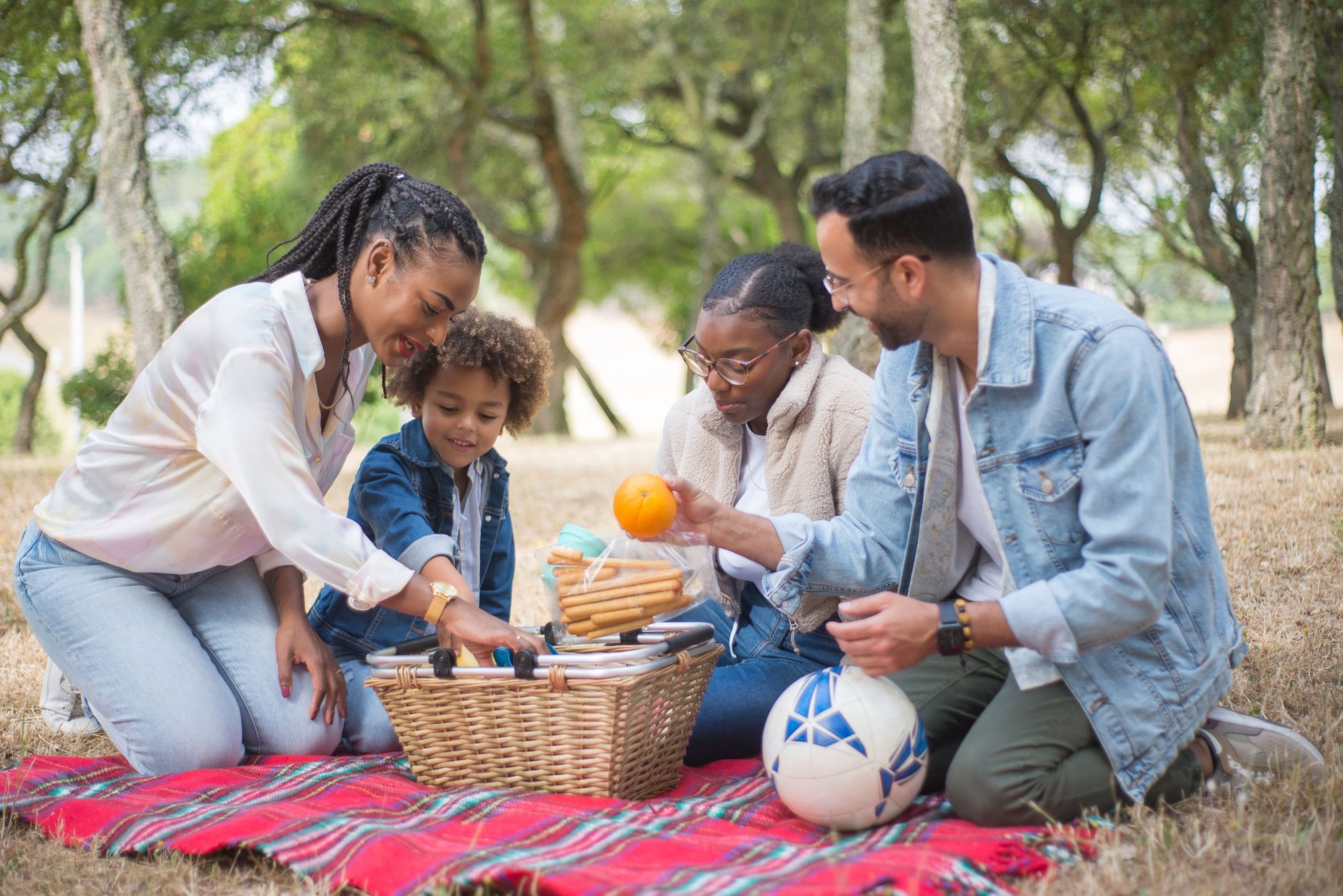A joyful family enjoying a picnic outdoors with snacks and a ball, surrounded by trees.