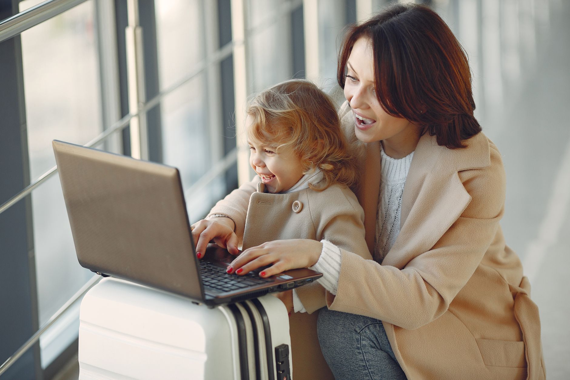 Cheerful stylish woman with suitcase hugging adorable laughing daughter and browsing laptop together while waiting for flight in airport hal