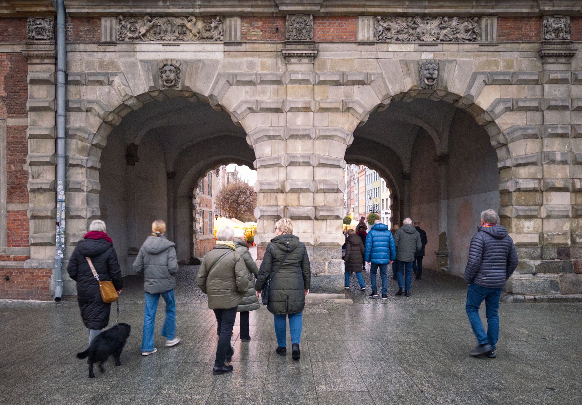 Tourists walk through a historic stone gate in Gdańsk, Poland, during a cool day.